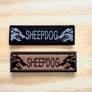 May include: Two rectangular embroidered patches. One is black with gray text and a gray graphic of a wolf-like creature. The text reads "SHEEPDOG". The other patch is brown with brown text and a brown graphic of a wolf-like creature. The text reads "SHEEPDOG".