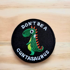 May include: A round, black embroidered patch featuring a green dinosaur with orange spikes. The text "DON'T BE A CUNTASAURUS" is embroidered in white around the dinosaur. The patch is on a light brown wooden surface.