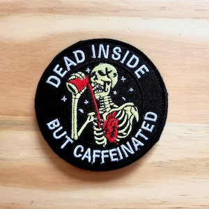 May include: Round black patch with the text "DEAD INSIDE BUT CAFFEINATED" surrounding a skeleton holding a red coffee cup. The skeleton has a red heart and yellow bones. The patch is on a light brown wooden surface.