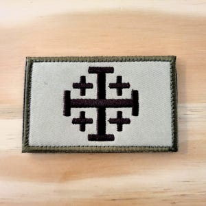 May include: A rectangular embroidered patch featuring a dark brown Jerusalem cross centered on a light gray background. The patch is finished with a dark olive green stitched border, creating a clean and defined edge for this fabric emblem.