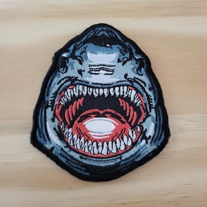 May include: Embroidered patch featuring a shark's open mouth with sharp teeth. The patch has a black border, with shades of blue and gray for the shark's head and a red interior mouth. The teeth are white.