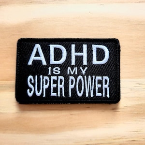May include: Black rectangular patch with white embroidered text that reads "ADHD IS MY SUPER POWER". The patch is set against a light brown wooden surface.