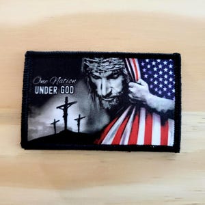 May include: A rectangular black patch featuring a grayscale image of Jesus wearing a crown of thorns and holding the American flag. The text "One Nation UNDER GOD" is printed on the patch.