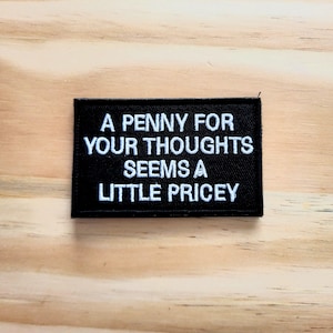 May include: A black rectangular patch with white embroidered text that reads "A PENNY FOR YOUR THOUGHTS SEEMS A LITTLE PRICE". The patch is placed on a light brown wooden surface.