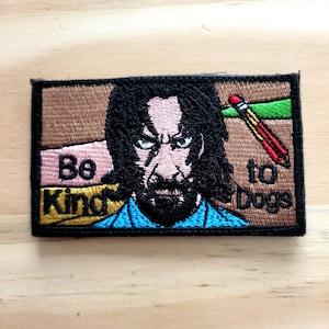 May include: Embroidered patch featuring a stern-faced character with long black hair and a beard. The patch includes the text "Be Kind to Dogs" with a pencil graphic. The patch has a brown, pink, and green color scheme.