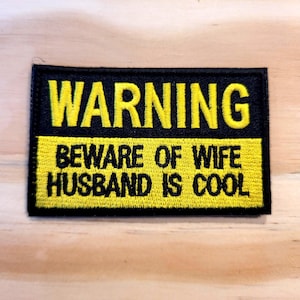 May include: A rectangular embroidered patch with a black background and yellow border. The top section features the word WARNING in yellow text. The bottom section has a yellow background with the black text BEWARE OF WIFE HUSBAND IS COOL.