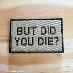 May include: Tan rectangular patch with the embroidered text "BUT DID YOU DIE?" in dark brown. The patch has a dark brown border and is set against a light wood grain background.