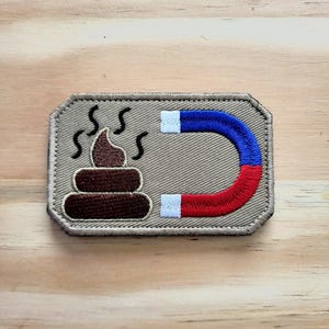 May include: Embroidered patch featuring a brown pile of poop with steam rising, next to a horseshoe magnet in red, blue, and white. The patch has a tan border and is rectangular in shape.