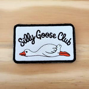 May include: A rectangular patch with a black border and the words "Silly Goose Club" in black script. A white goose with an orange beak and feet is lying down on the patch.