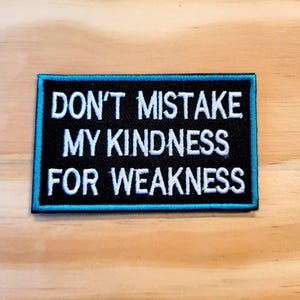 May include: A rectangular embroidered patch with a black background and a bright blue border. The patch features the white embroidered text DON'T MISTAKE MY KINDNESS FOR WEAKNESS arranged in three lines across the center of the piece.