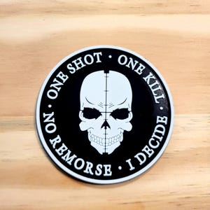 May include: A circular sticker featuring a white skull graphic centered on a black background. A vertical crosshair line runs down the center of the skull. The text "ONE SHOT ONE KILL NO REMORSE I DECIDE" is printed in white around the border.