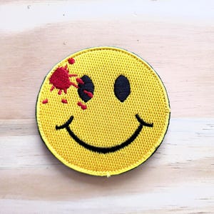 May include: Embroidered yellow smiley face patch with black eyes and a stitched black smile. The patch has a red blood splatter design on the left side. The patch is round with a black border.