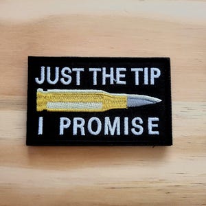 May include: Black rectangular patch with the text "JUST THE TIP I PROMISE" in white. A yellow and silver bullet graphic is centered on the patch. The patch is on a light brown wooden surface.