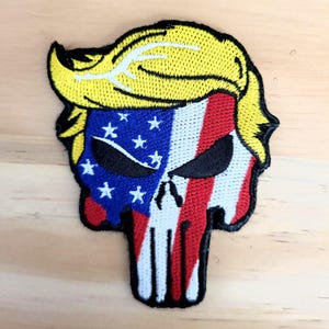 May include: Embroidered patch featuring a skull design with a yellow hairstyle, a blue section with white stars, and red and white stripes. The patch has a black border and is likely for clothing or accessories.