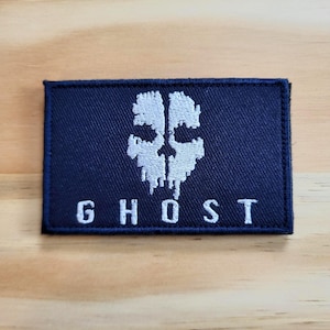 May include: A navy blue rectangular patch with a white skull design and the word "GHOST" in white block letters. The patch is likely made of fabric and has a stitched border.