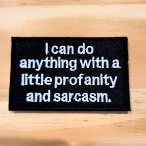 May include: Black rectangular patch with white embroidered text: "I can do anything with a little profanity and sarcasm." The patch is on a light brown wooden surface.