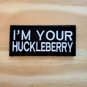 May include: Black rectangular patch with white embroidered text that reads "I'M YOUR HUCKLEBERRY". The patch is likely for clothing or accessories, and is displayed on a light wood surface.