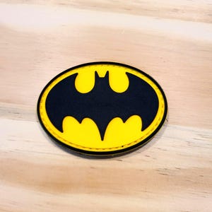 May include: An oval-shaped patch featuring a black bat silhouette centered on a bright yellow background. The patch has a thick black border with visible stitching along the edge, creating a distinct contrast between the yellow and black colors.