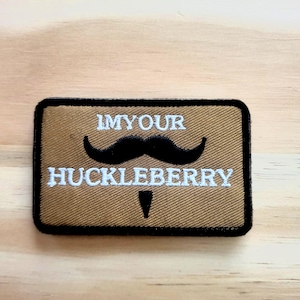 May include: A rectangular patch with a brown background and black border. The patch features a black mustache and the white text "IMYOUR HUCKLEBERRY". The patch is likely an accessory for clothing or bags.