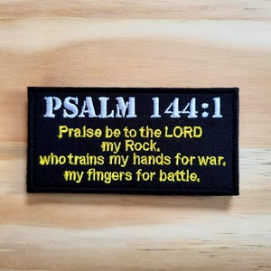 May include: Black rectangular patch with white and yellow embroidered text. The text reads "PSALM 144:1" at the top, followed by a quote from the Bible. The patch is on a light brown wooden surface.