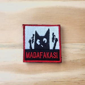 May include: Embroidered patch featuring a black cat silhouette holding two guns. The cat has wide eyes and is set against a white background with a red border. The text "MADAF*KAS!" is displayed in red letters.