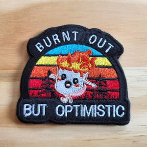 May include: Embroidered patch with the words "BURNT OUT BUT OPTIMISTIC" in white lettering on a black background. The patch features a cartoon character with flames on its head, set against a sunset-colored backdrop.