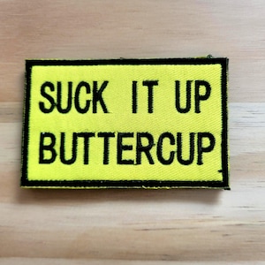 May include: A rectangular patch with the text "SUCK IT UP BUTTERCUP" embroidered in black on a bright yellow background. The patch has a black border and is likely designed to be sewn or ironed onto clothing or accessories.