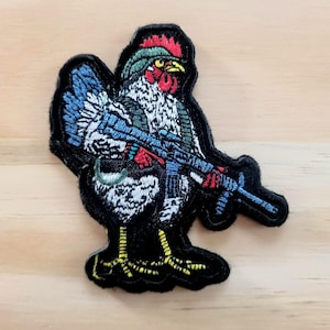 May include: Embroidered patch featuring a rooster holding a blue rifle. The rooster has a red comb, green head covering, and blue and white feathers. The patch has a black border and is set against a light wood grain background.