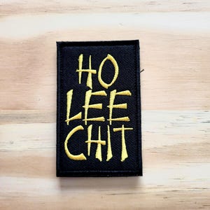 May include: Black rectangular patch with the embroidered text "HO LEE CHIT" in yellow. The patch is likely made of fabric and is set against a light wood grain background.