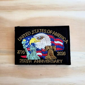 May include: A black patch with gold text reading "UNITED STATES OF AMERICA" and "250TH ANNIVERSARY". The patch features an eagle, the Statue of Liberty, and a waving American flag. Dates 1776 and 2026 are also included.