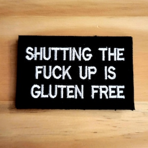 May include: Black rectangular patch with white embroidered text that reads: "SHUTTING THE FUCK UP IS GLUTEN FREE". The patch is on a light brown wooden surface.