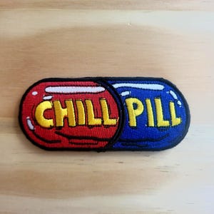 May include: A colorful patch in the shape of a pill, divided into red and blue sections. The word "CHILL" is in yellow letters on the red side, and "PILL" is in yellow on the blue side. The patch has a black border.