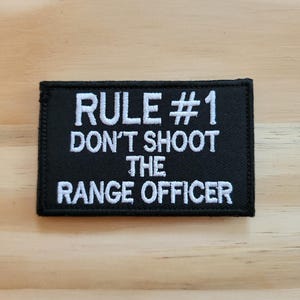May include: Black rectangular patch with white embroidered text. The text reads "RULE #1 DON'T SHOOT THE RANGE OFFICER". The patch has a black border and is set against a light wood grain background.