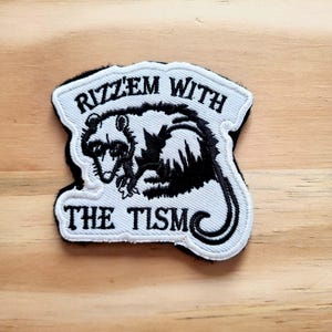 May include: Embroidered patch featuring a black and white raccoon illustration. The patch has the text "Rizz'em With The Tism" in a stylized font, with a black border. The patch is on a light wood surface.