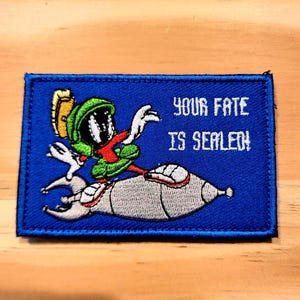 May include: Embroidered patch featuring Marvin the Martian riding a rocket. The patch is blue with white text that reads "YOUR FATE IS SEALED!". Marvin is depicted in his signature green, red, and white attire.