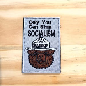 May include: A rectangular embroidered patch with a light gray background featuring a brown bear wearing a park ranger style hat with the word PATRIOT on the brim. The text above the bear reads Only You Can Stop SOCIALISM in black lettering.