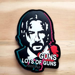 May include: A black and white graphic of a man's face with the words "GUNS LOTS OF GUNS PRINCIPLE" in white and red text. The image has a black border with teal and pink accents. The image is on a light brown wooden surface.