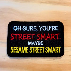 May include: A rectangular black embroidered patch with rounded corners featuring white, red, and yellow text. The text reads: OH SURE, YOU'RE STREET SMART. MAYBE SESAME STREET SMART. The patch has a thick black border and is stitched with high-contrast thread.