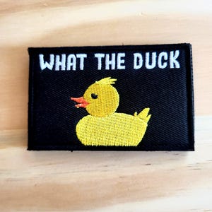 May include: Embroidered black patch with the phrase "WHAT THE DUCK" in white letters above a yellow duck with an orange beak. The duck is embroidered with yellow thread. The patch is rectangular.