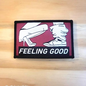 May include: A rectangular patch with a black border and a maroon background. The patch features a stylized illustration of legs, one in high heels and the other in sneakers. The words "FEELING GOOD" are printed in white.