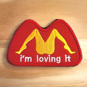 May include: Embroidered patch with a red background and a yellow "M" shape formed by legs. The text "I'm loving it" is in white letters below the graphic. The patch has a black border and is on a wooden surface.