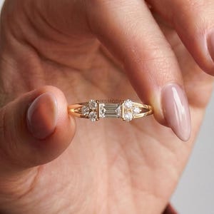 Puede incluir: Un anillo de oro con un diamante central de corte baguette flanqueado por dos diamantes más pequeños de corte redondo a cada lado. La banda es fina y pulida, mostrando una disposición simétrica de cinco piedras claras engastadas horizontalmente.