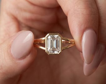 May include: A gold ring featuring a central emerald-cut clear gemstone set in a bezel setting. The band is designed with a split shank that tapers toward the center stone, creating a sleek and refined appearance with a polished metallic finish.