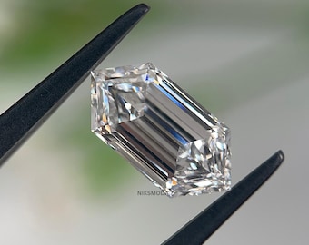 May include: A clear, emerald-cut diamond held by black tweezers. The diamond has a rectangular shape with cut corners and multiple facets, reflecting light. The background is blurred, with a hint of green.