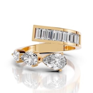 Può includere: Un anello aperto in oro con una fila di pietre taglio baguette su un'estremità e tre pietre taglio a goccia sull'altra. La fascia è in oro lucido e la parola NIK SMODA è stampata in piccole lettere nere sulla superficie interna dell'anello.