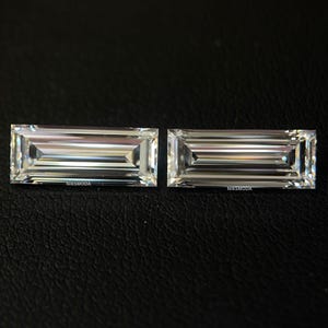 Puede incluir: Dos gemas rectangulares y transparentes con talla brillante, colocadas una al lado de la otra. Las gemas tienen una serie de facetas paralelas, creando un efecto brillante. El nombre de la marca "NIKSMODA" es visible en cada piedra. El fondo es oscuro.