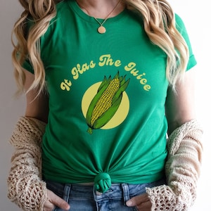 Può includere: Una maglietta verde con una grafica di mais giallo e verde su un'orecchia e il testo "It Has The Juice" in un font retrò.