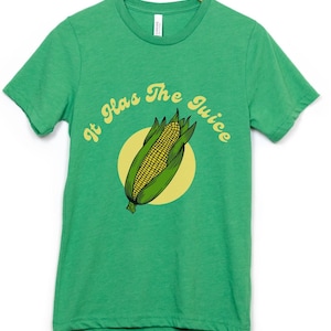 Può includere: Maglietta verde con un grafico di mais giallo e verde su un'orecchia e il testo "It Has The Juice" in un font retrò.