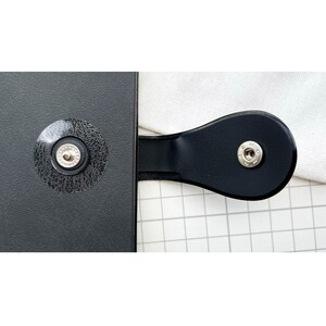 May include: Close-up of a black leather-bound notebook with two silver metal snaps. The notebook is closed and the snaps are visible.
