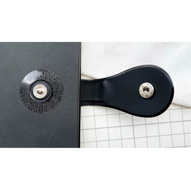 May include: Close-up of a black leather-bound notebook with two silver metal snaps. The notebook is closed and the snaps are visible.